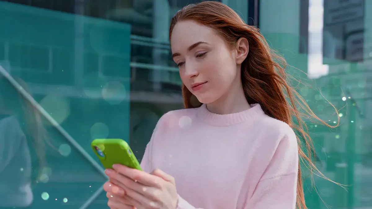 A young woman outdoors in a pink jumper looks at her mobile phone with an enigmatic expression. This is Virgin Media's guide to freeing up phone storage space.