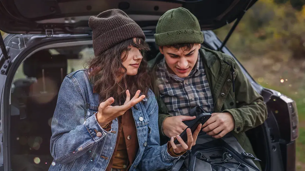 A woman and a man in outdoor clothing rest on a car with an open boot while looking at their mobile phones. This is Virgin Media's guide to freeing up phone storage space.