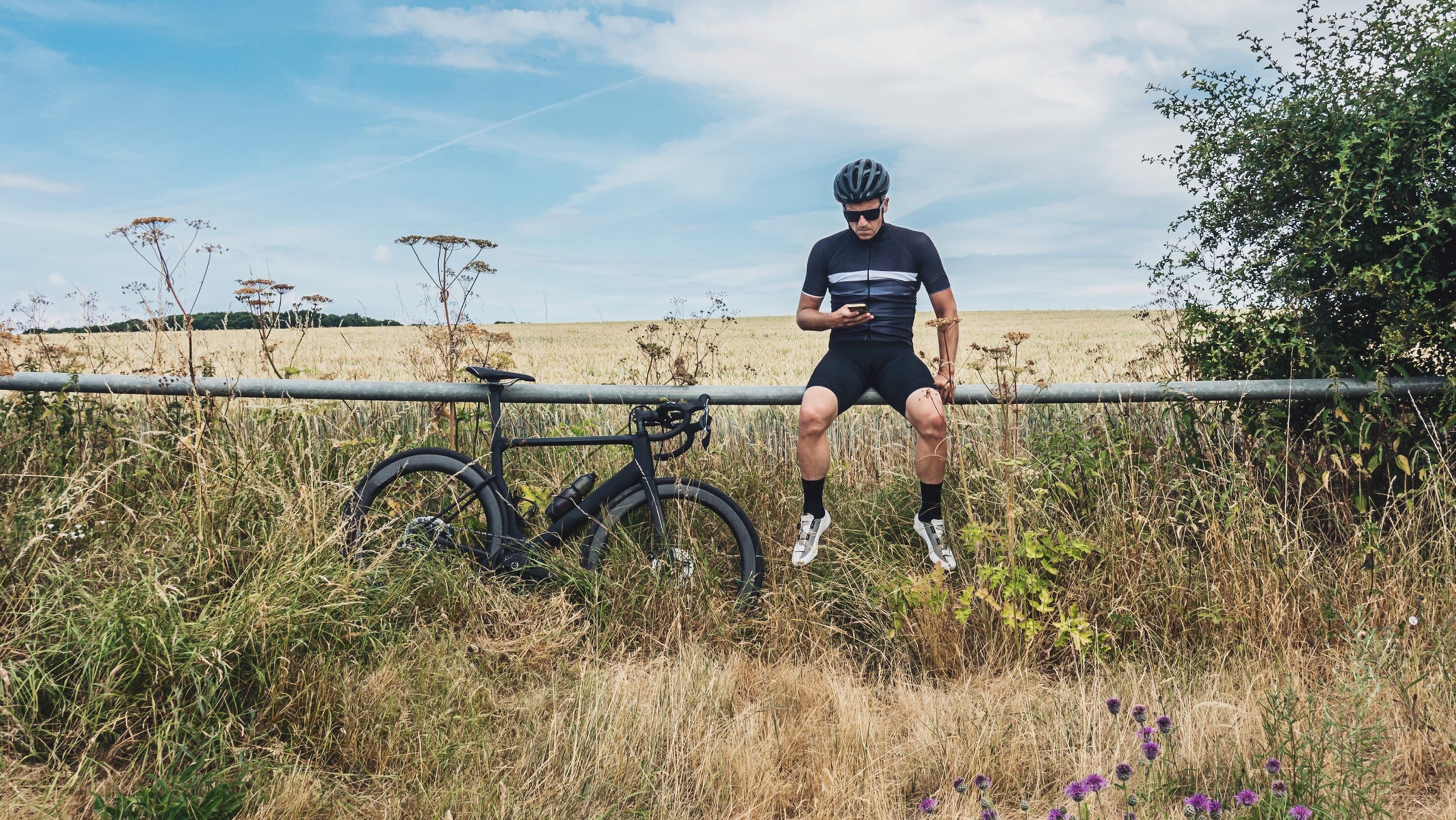 Cyclist in black gear sits on a rural fence, checking his phone. A bicycle rests nearby amid tall grass and wildflowers under a blue sky.
