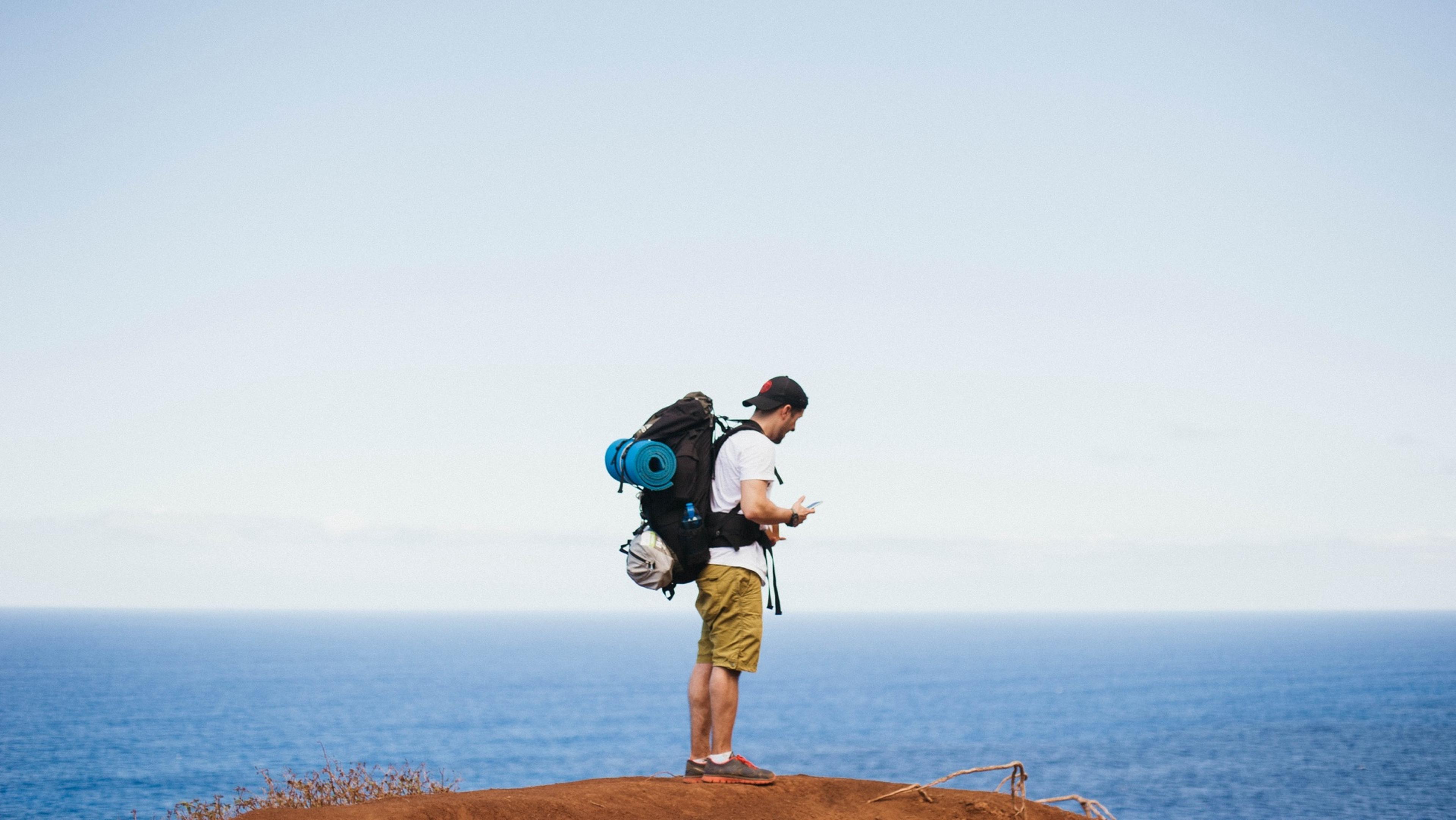 A hiker with a backpack stands on a cliff overlooking the ocean, checking his phone under a clear blue sky.