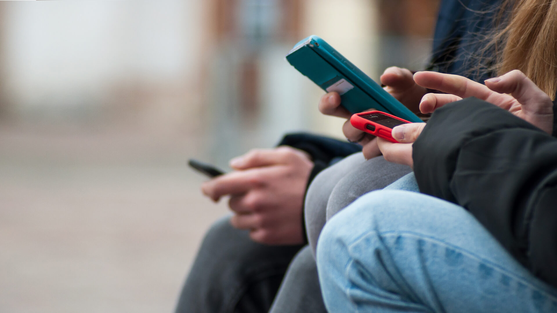 Close-up of three people using smartphones outdoors, focusing on hands holding devices with colorful cases.