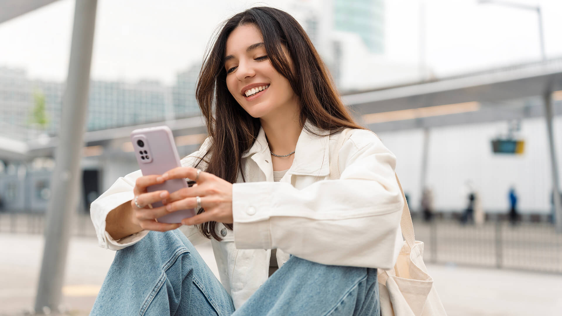 Smiling woman in a white jacket and jeans, sitting outdoors, looking at her phone.