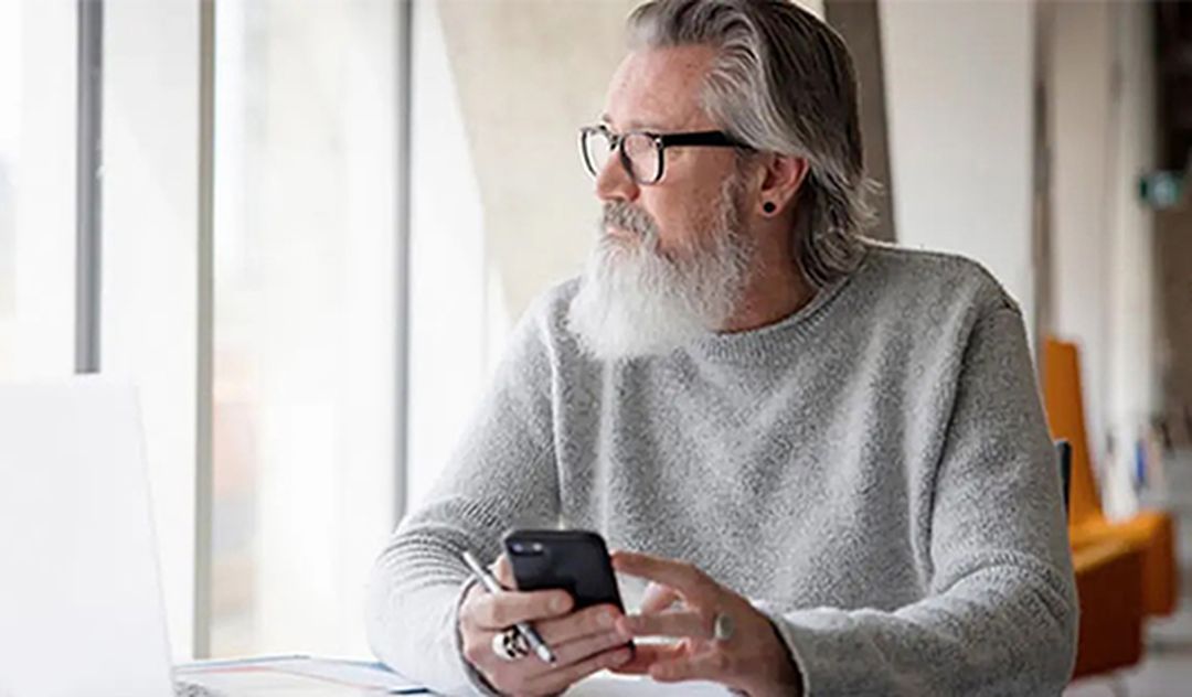 A bearded man with glasses sits at a table, holding a smartphone and looking out the window thoughtfully.