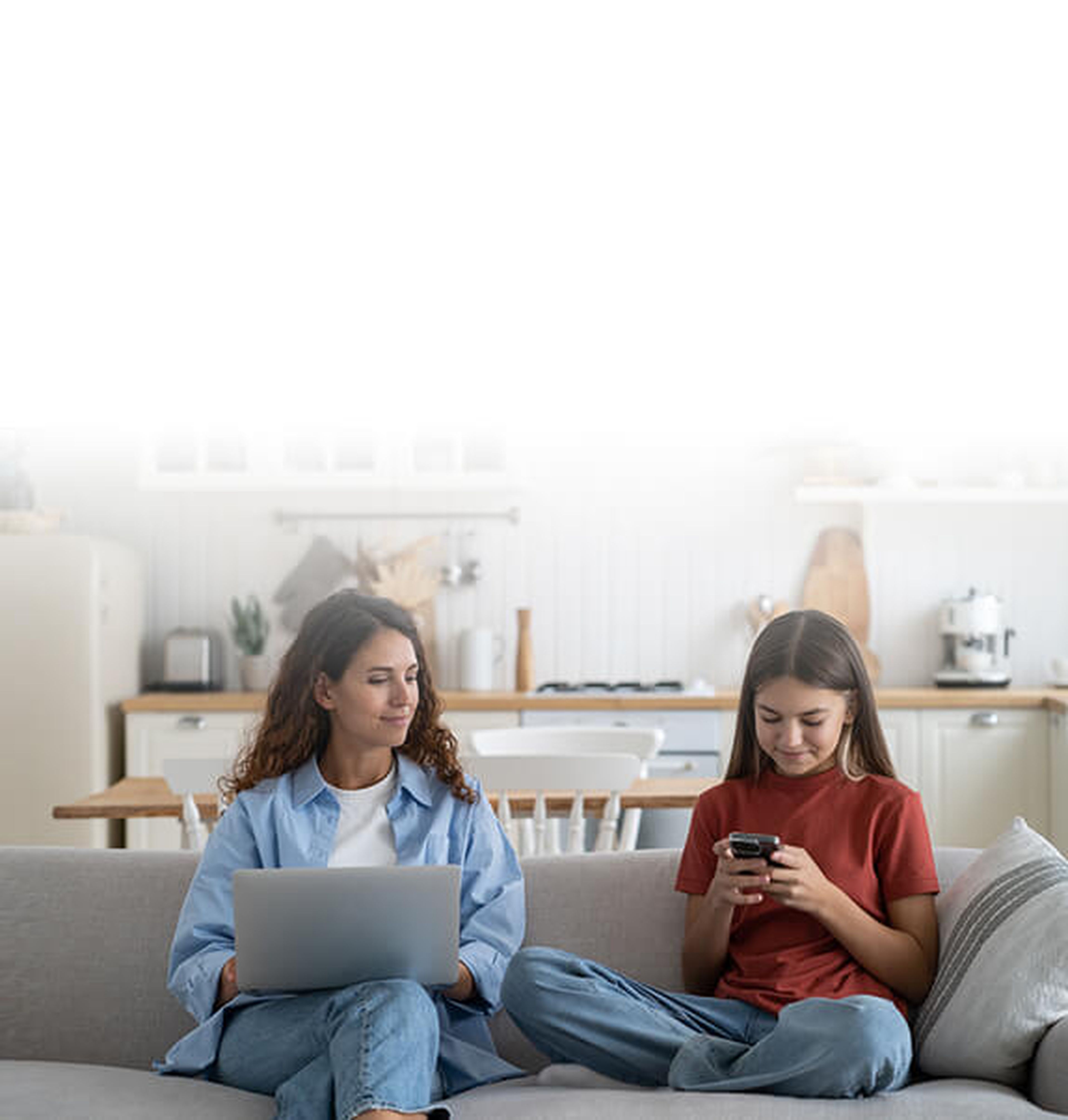 Mother using laptop and teen daughter using smartphone. They both sit on a sofa