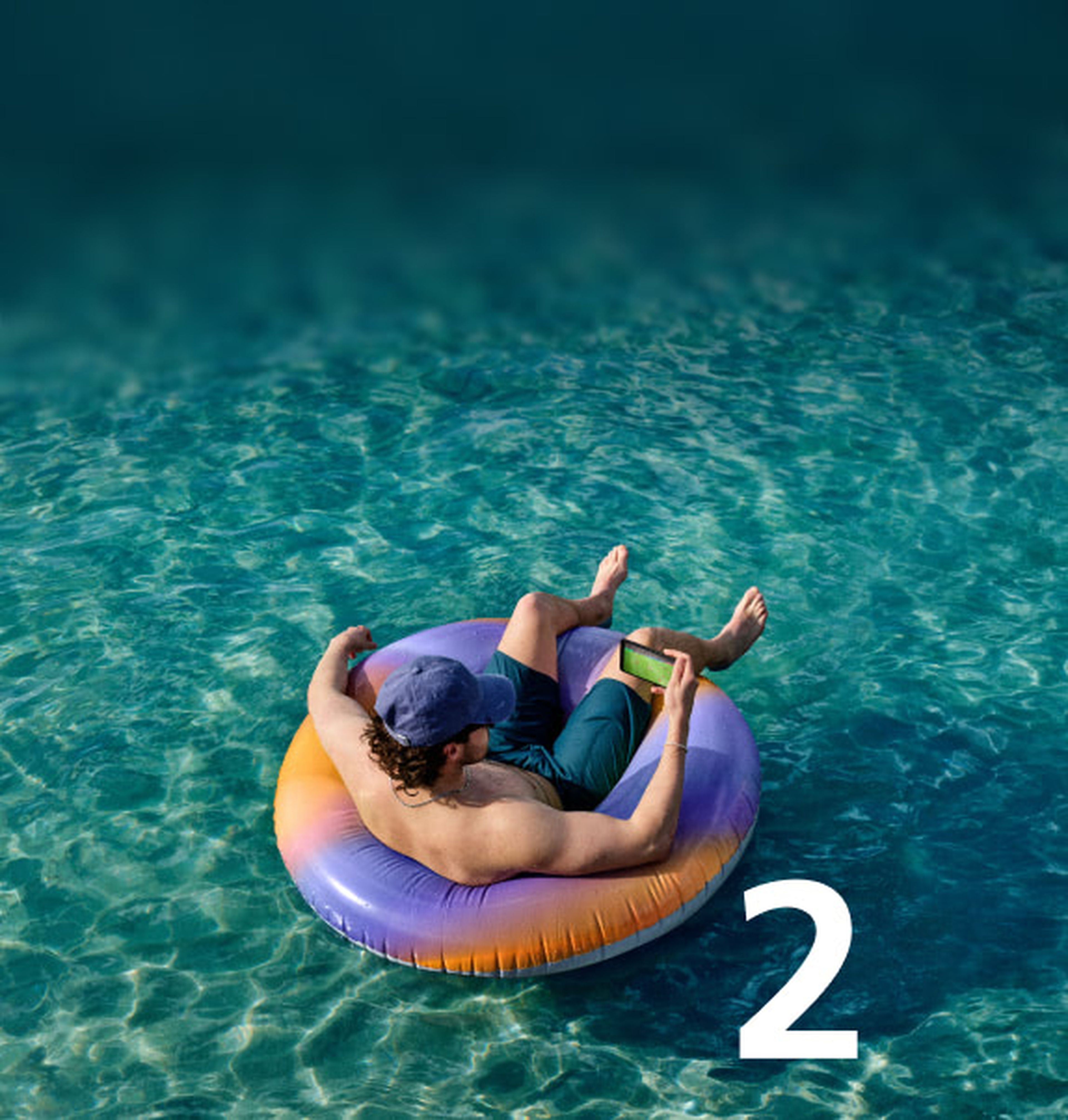 Person in a cap and shorts floating on an inflatable ring in clear blue water, holding a smartphone. Large number 2 on the water.