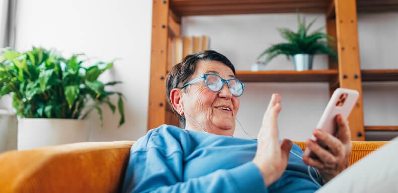 Elderly person with glasses smiles, wearing earphones, and wave while holding a smartphone, seated on a couch. There's greenery in the background.