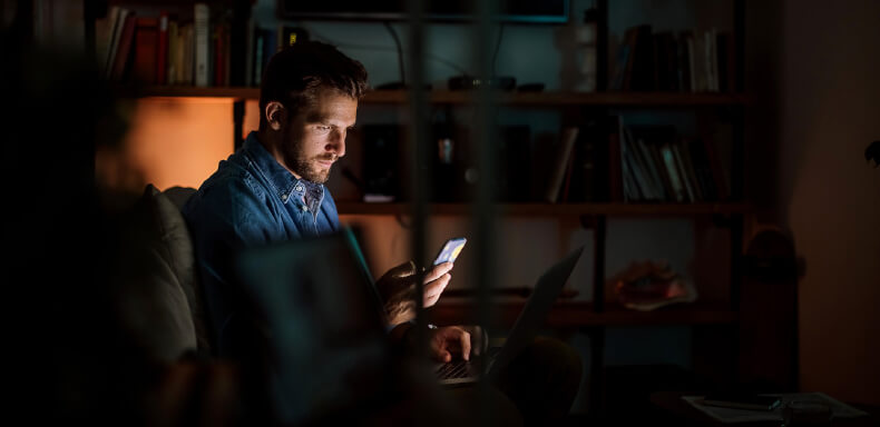 Man in a dimly lit room looking at a smartphone, with a laptop on his lap. Bookshelves are visible in the background.