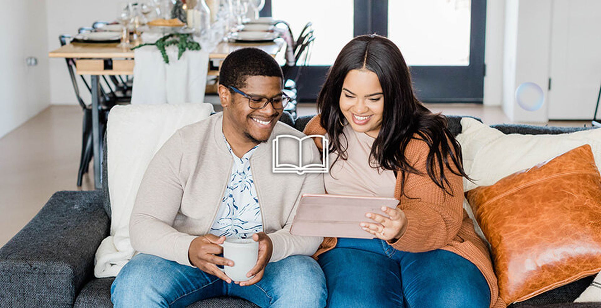 A smiling couple sits on a couch, looking at a tablet together, with a dining table set in the background.