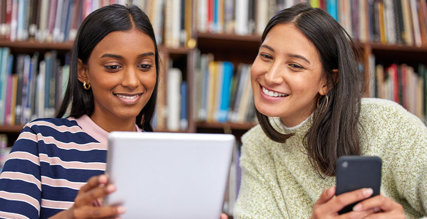 Two women smiling and looking at a tablet in a library, with bookshelves in the background.