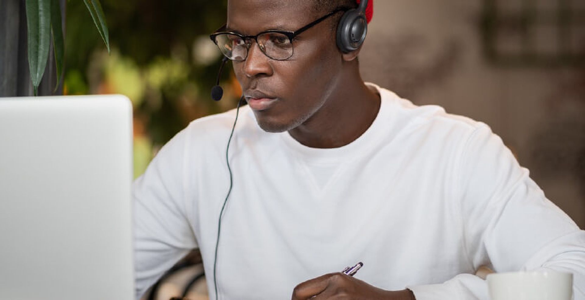 A person wearing glasses and a headset is working intently on a laptop, holding a pen, with a cup beside them.