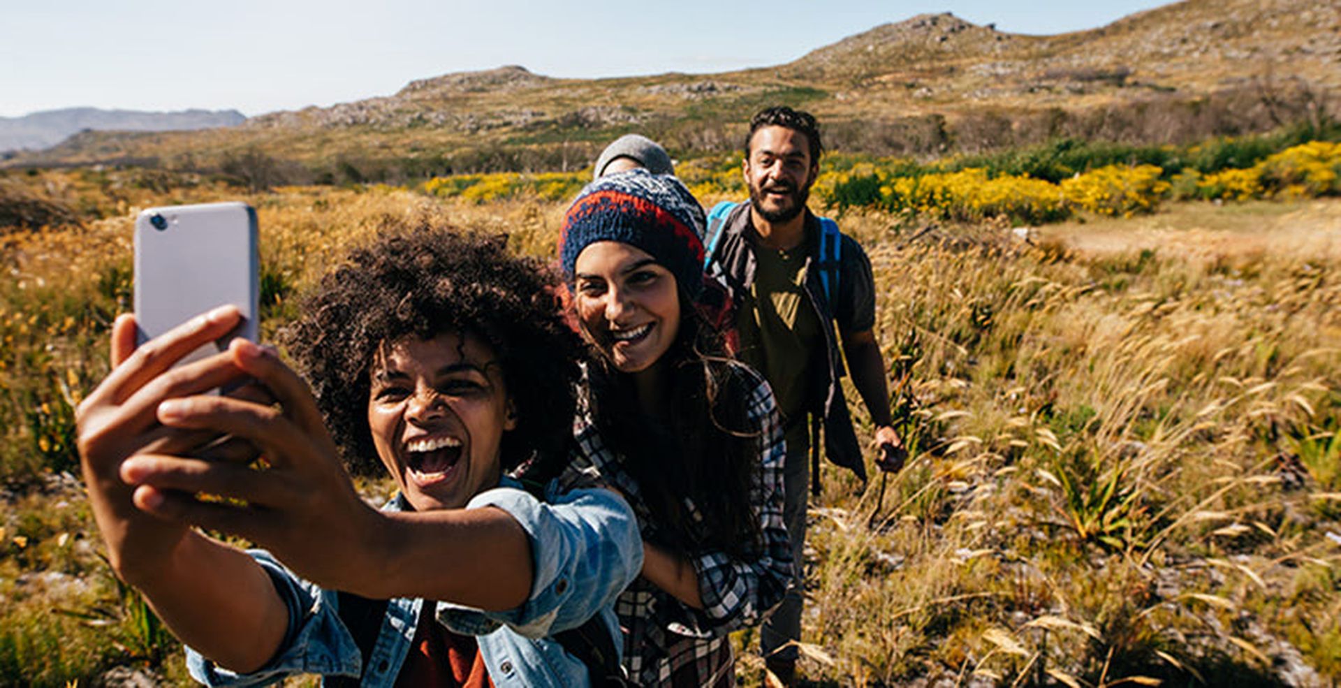 Three people smiling and taking a selfie while hiking in a grassy field, with mountains and yellow flowers in the background.