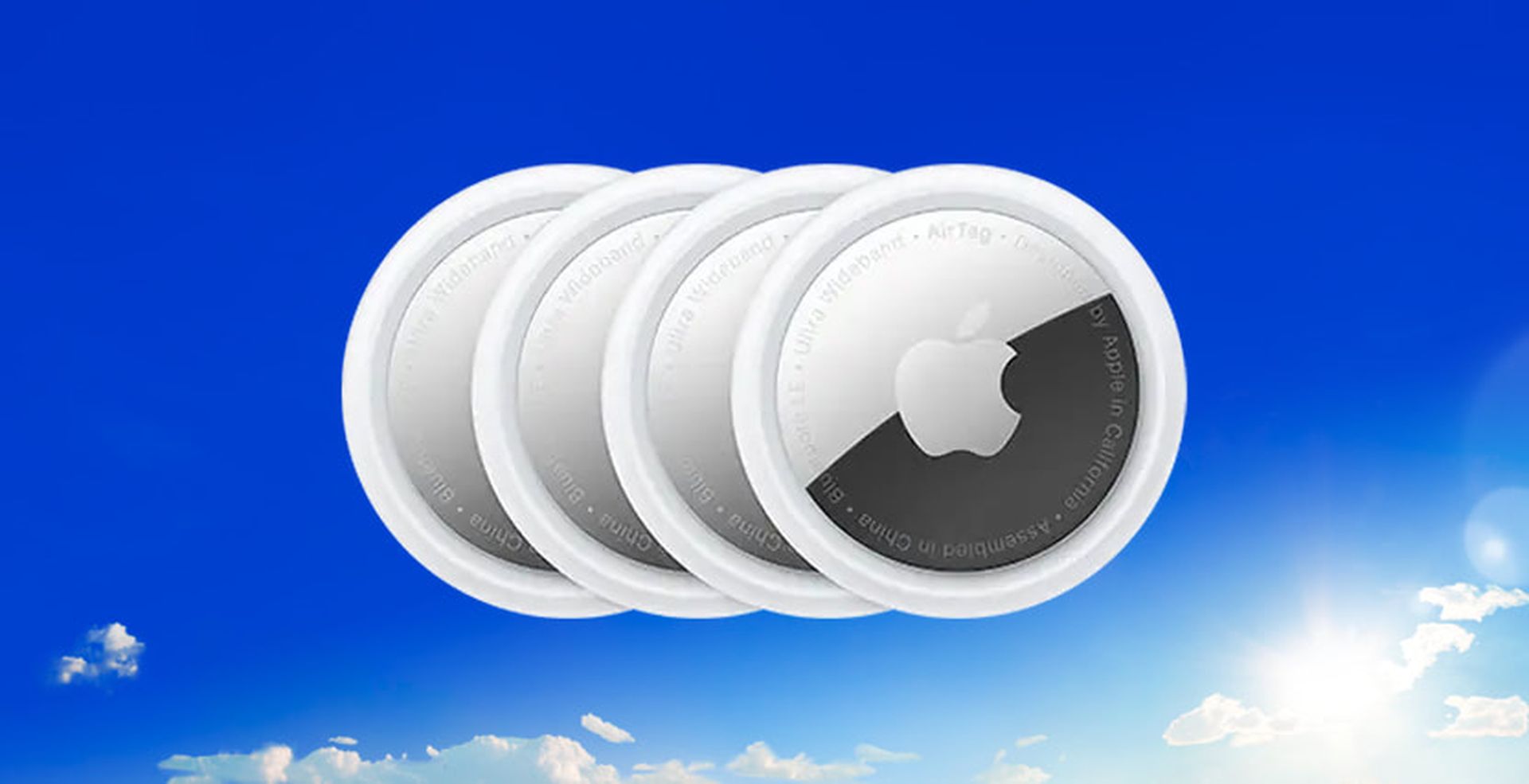 Four Apple AirTags aligned in a row against a bright blue sky with a few clouds and the sun shining.