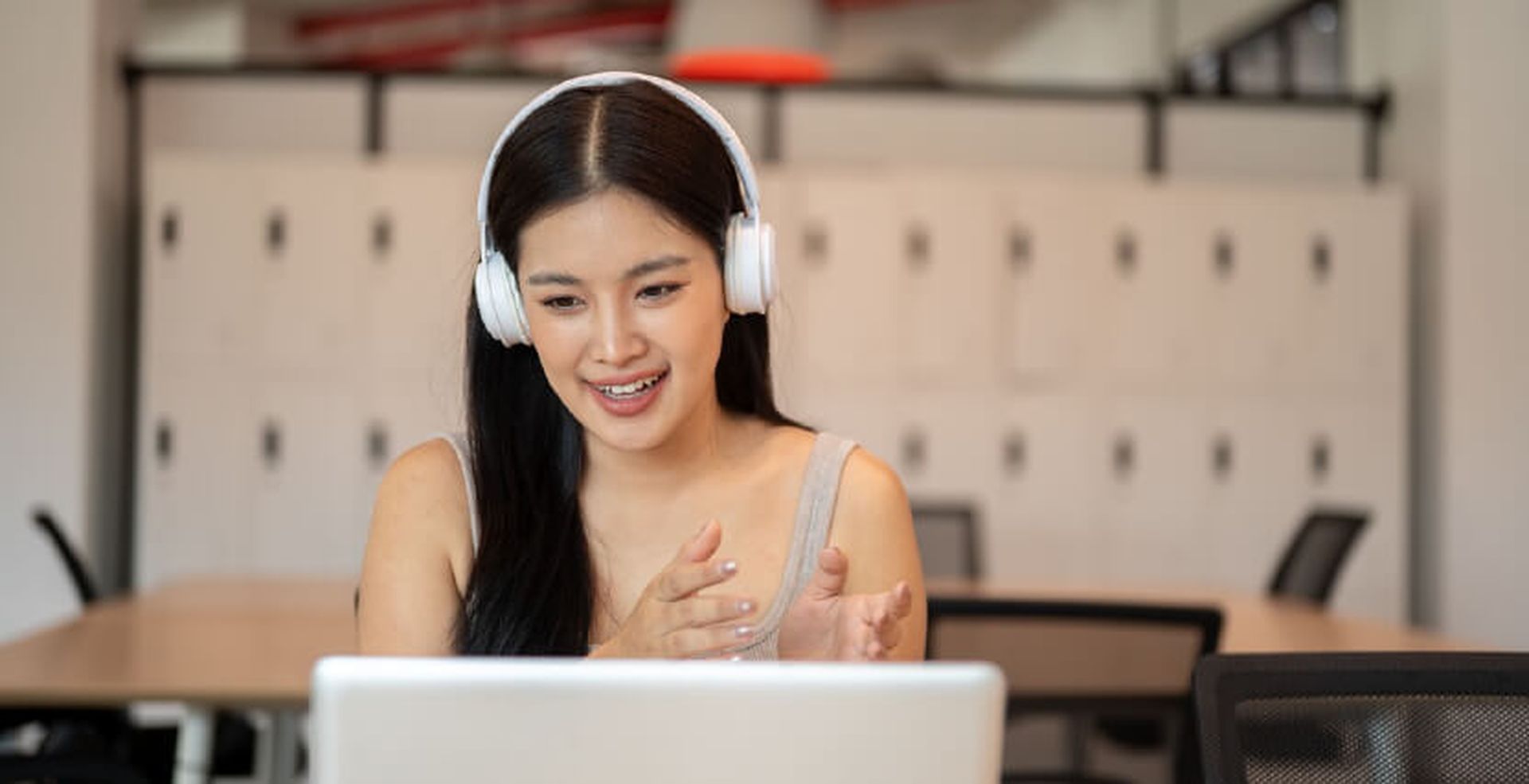 A woman with long hair wearing headphones claps while looking at a laptop in an office setting.
