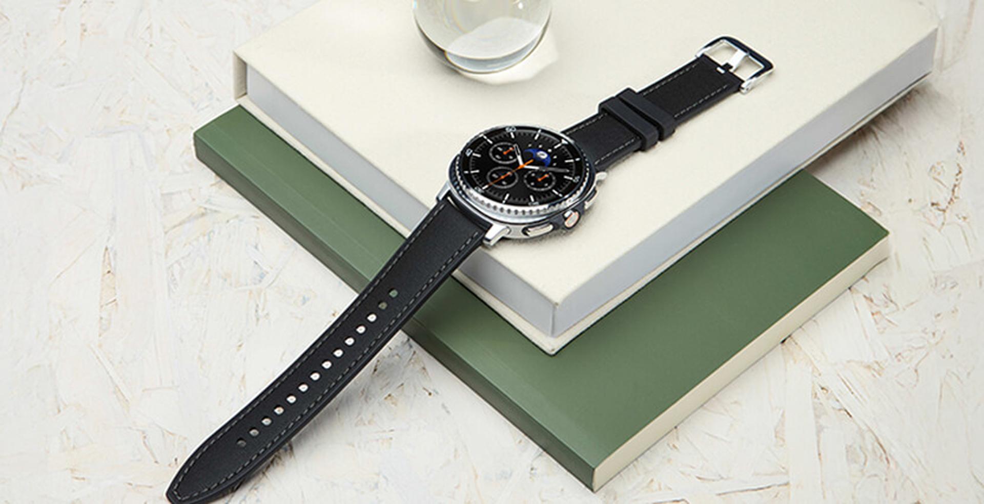 A black leather-strapped watch with a round, dark face rests on stacked books, with a small glass orb nearby.
