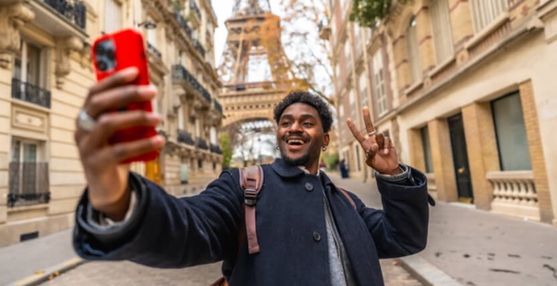 A person takes a selfie with a red phone on a Paris street, smiling and holding up a peace sign, with the Eiffel Tower in the background.