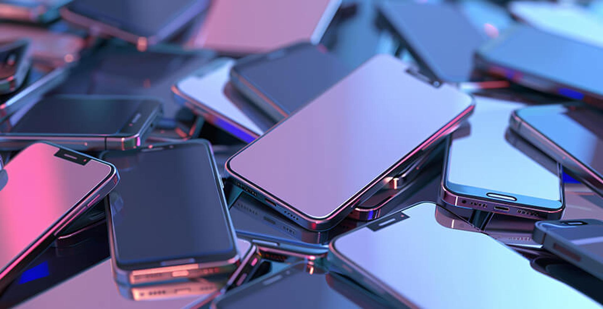 A pile of modern smartphones with metallic edges and reflective screens, illuminated by purple and blue lighting.