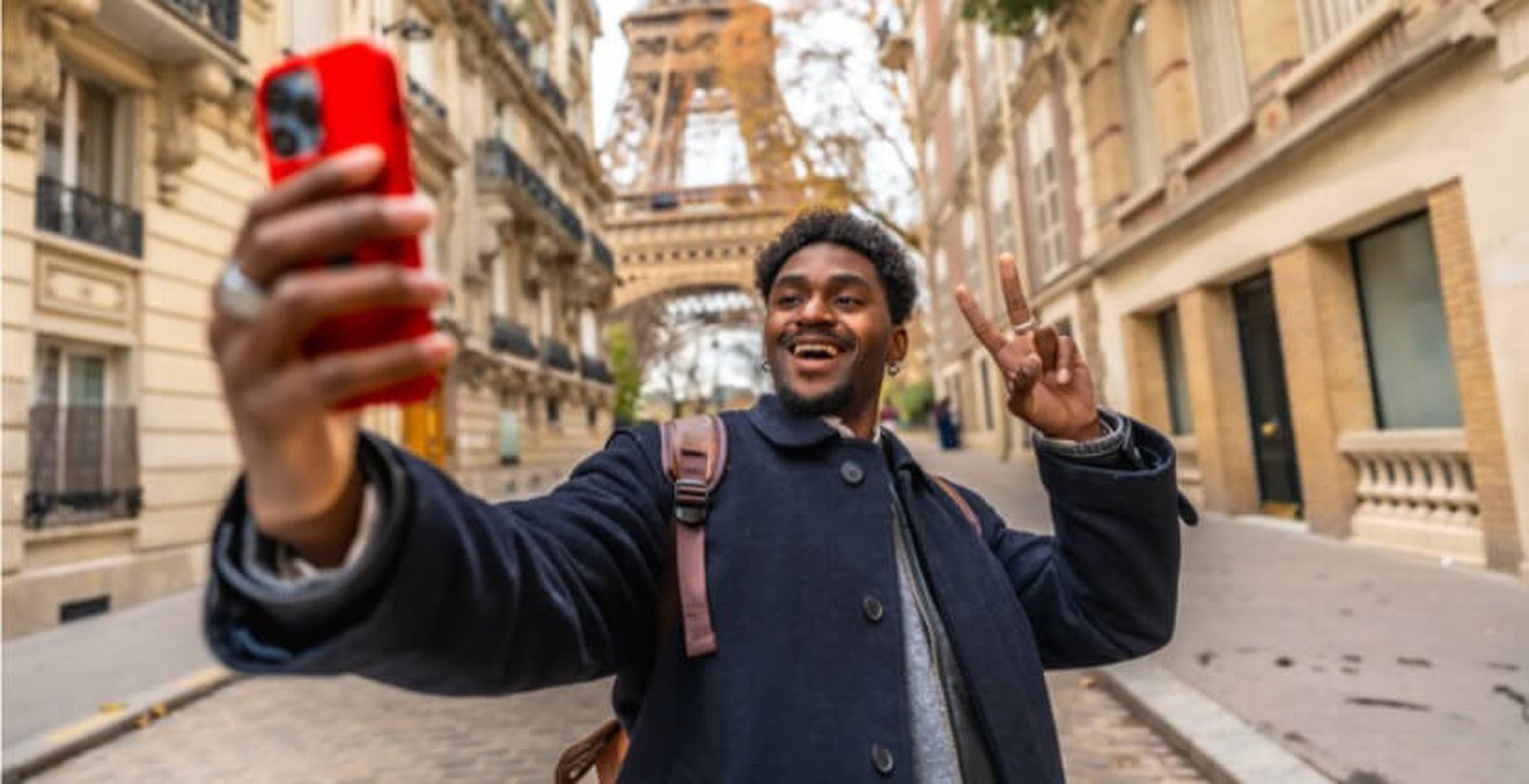 A man takes a selfie with a red phone in front of the Eiffel Tower, smiling and making a peace sign on a street lined with buildings.