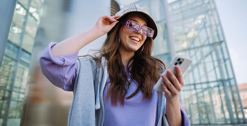 Woman in a purple outfit and sunglasses smiles while looking at her phone, standing in front of a modern glass building.