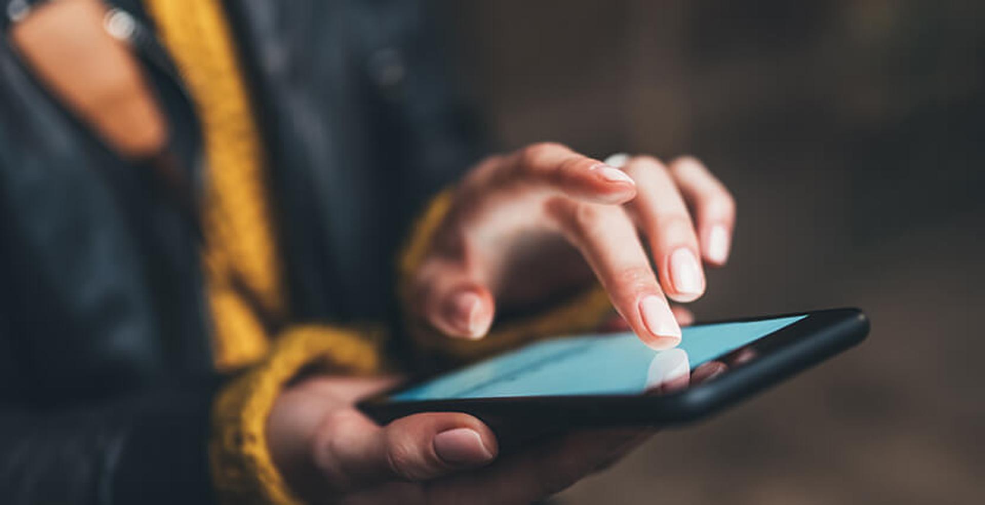 Close-up of a person wearing a yellow sweater and black jacket, using a black smartphone with one hand while touching the screen with the other.