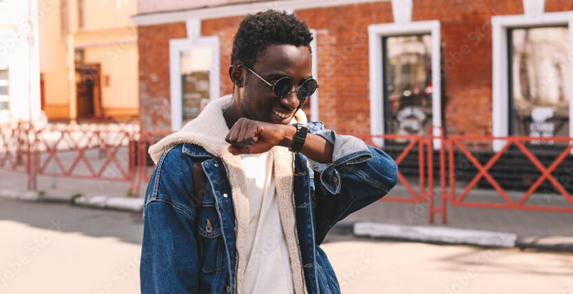 A person in sunglasses and a denim jacket checks their smartwatch, smiling, with a brick building and red fence in the background.