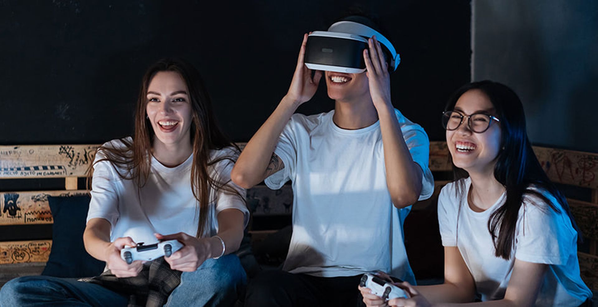 Three friends in white t-shirts enjoying a gaming session; two with controllers, one wearing a VR headset, all smiling and sitting on a couch.