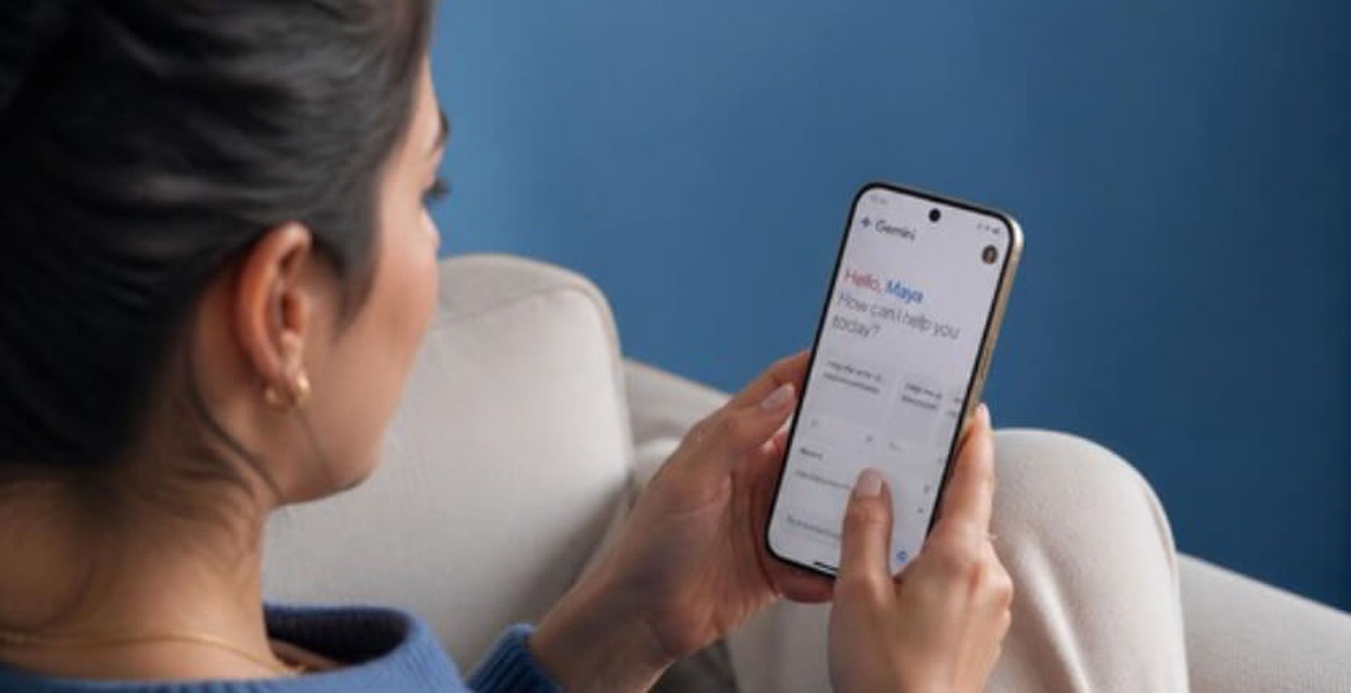 A woman sits on a couch holding a smartphone, interacting with a digital assistant on the screen, against a blue wall background.