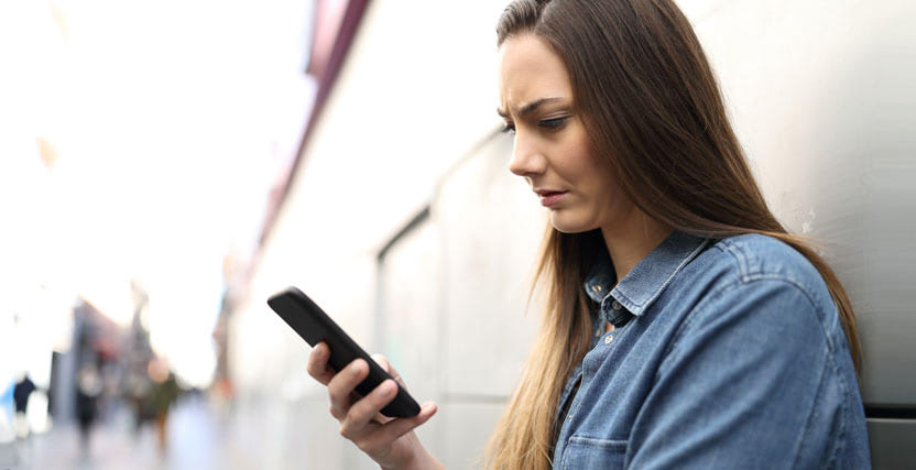 Woman in a denim shirt looks concerned while reading something on her smartphone, standing against an outdoor wall.