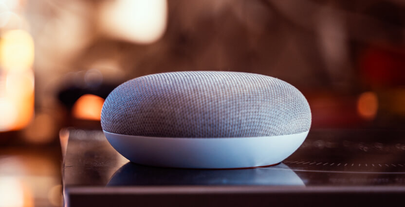 A smart speaker with a gray fabric top sits on a reflective surface, indoors. The background is blurred and warm-toned.