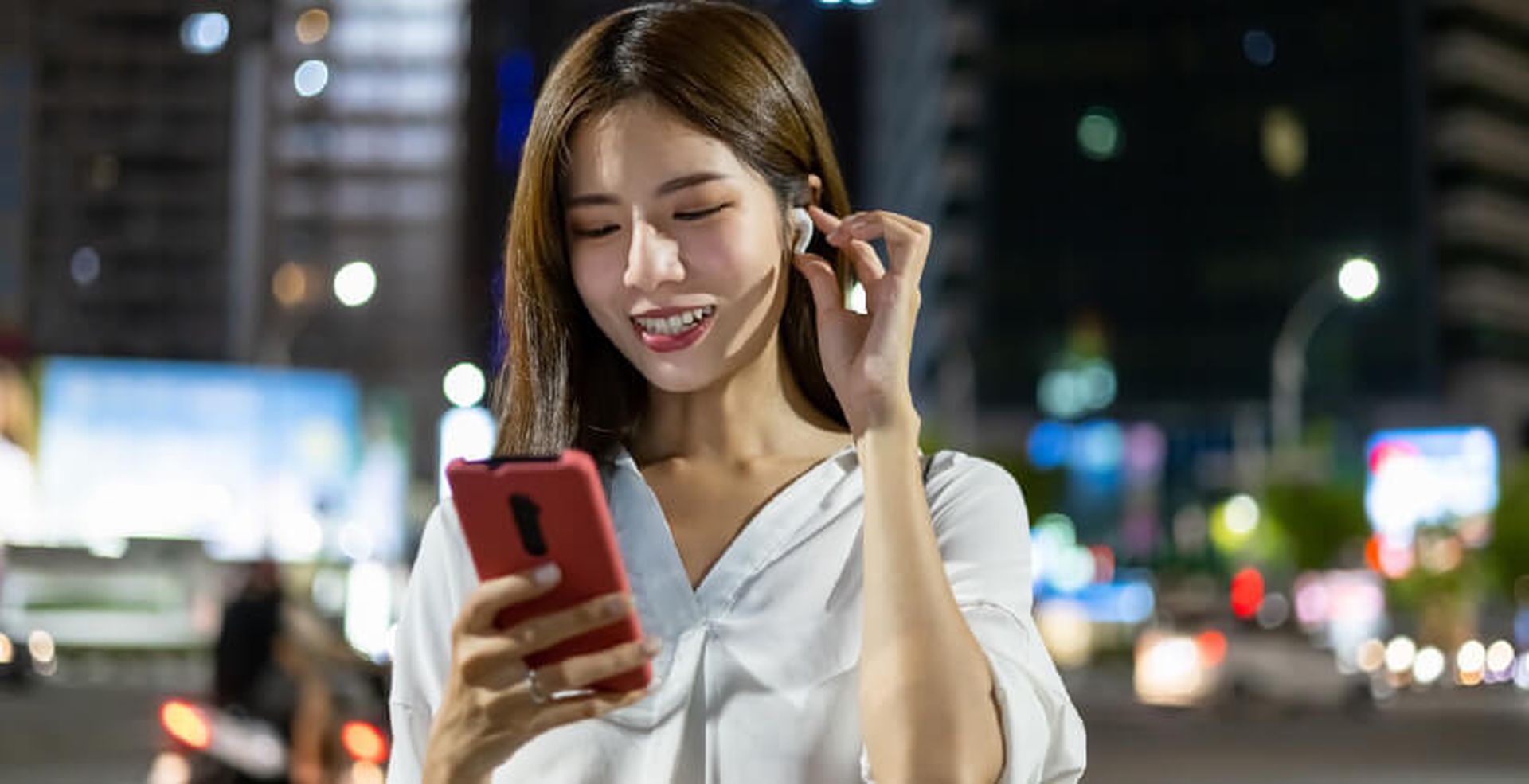 Woman smiling while looking at smartphone and adjusting earbuds, standing on a city street at night with blurred lights in the background.