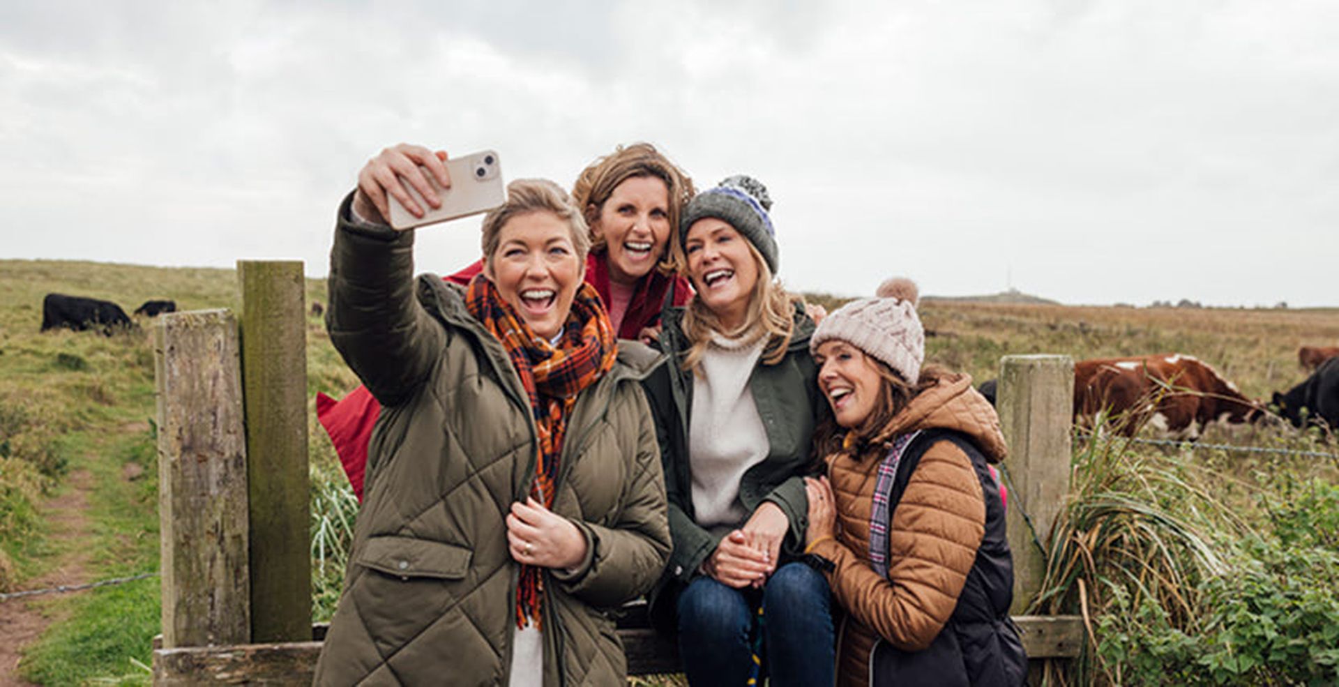 Four women dressed in warm clothing take a selfie while sitting on a wooden fence in a rural field with cows in the background.