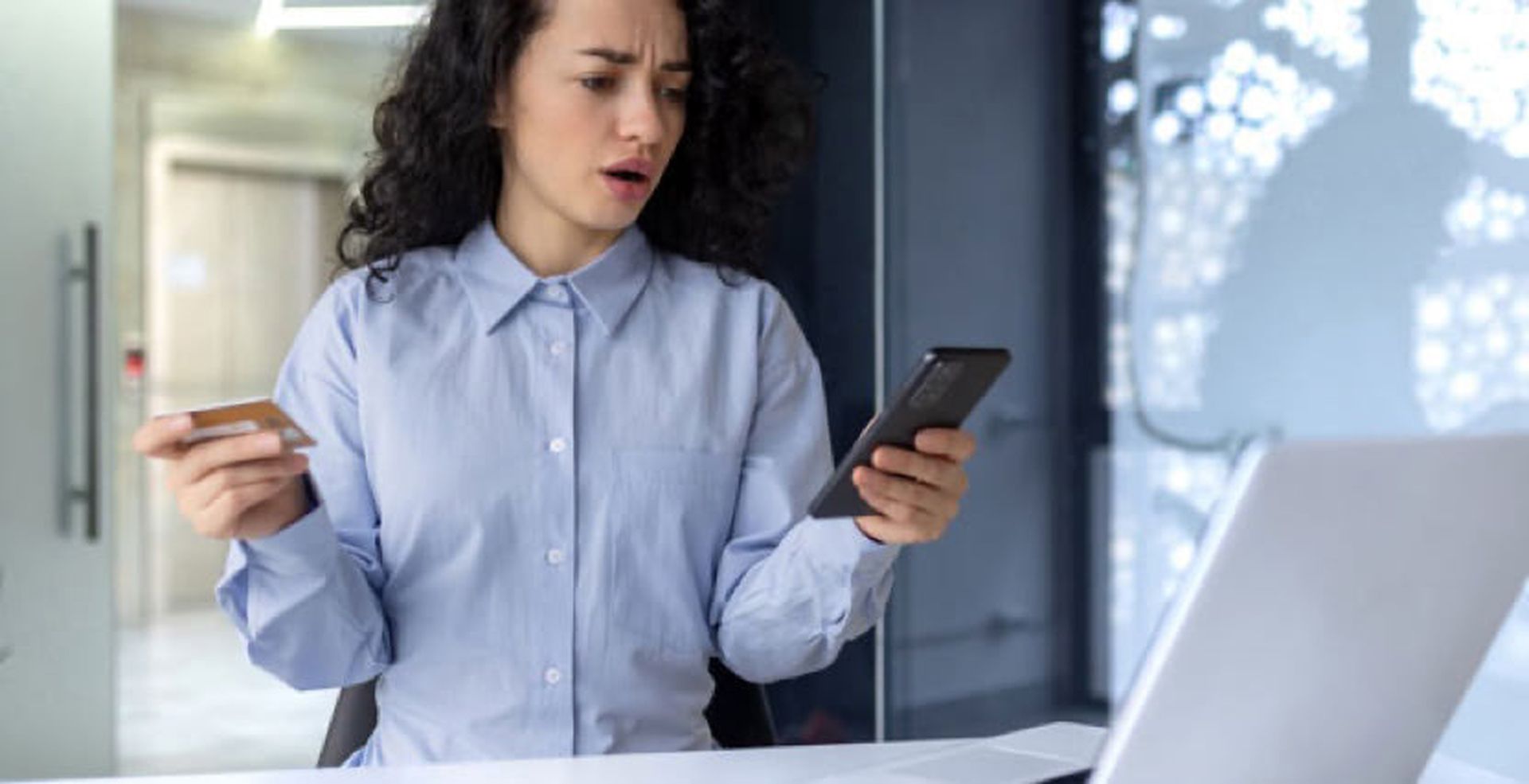 Woman with curly hair, wearing a blue shirt, looks surprised at her phone while holding a credit card, sitting at a desk with a laptop.