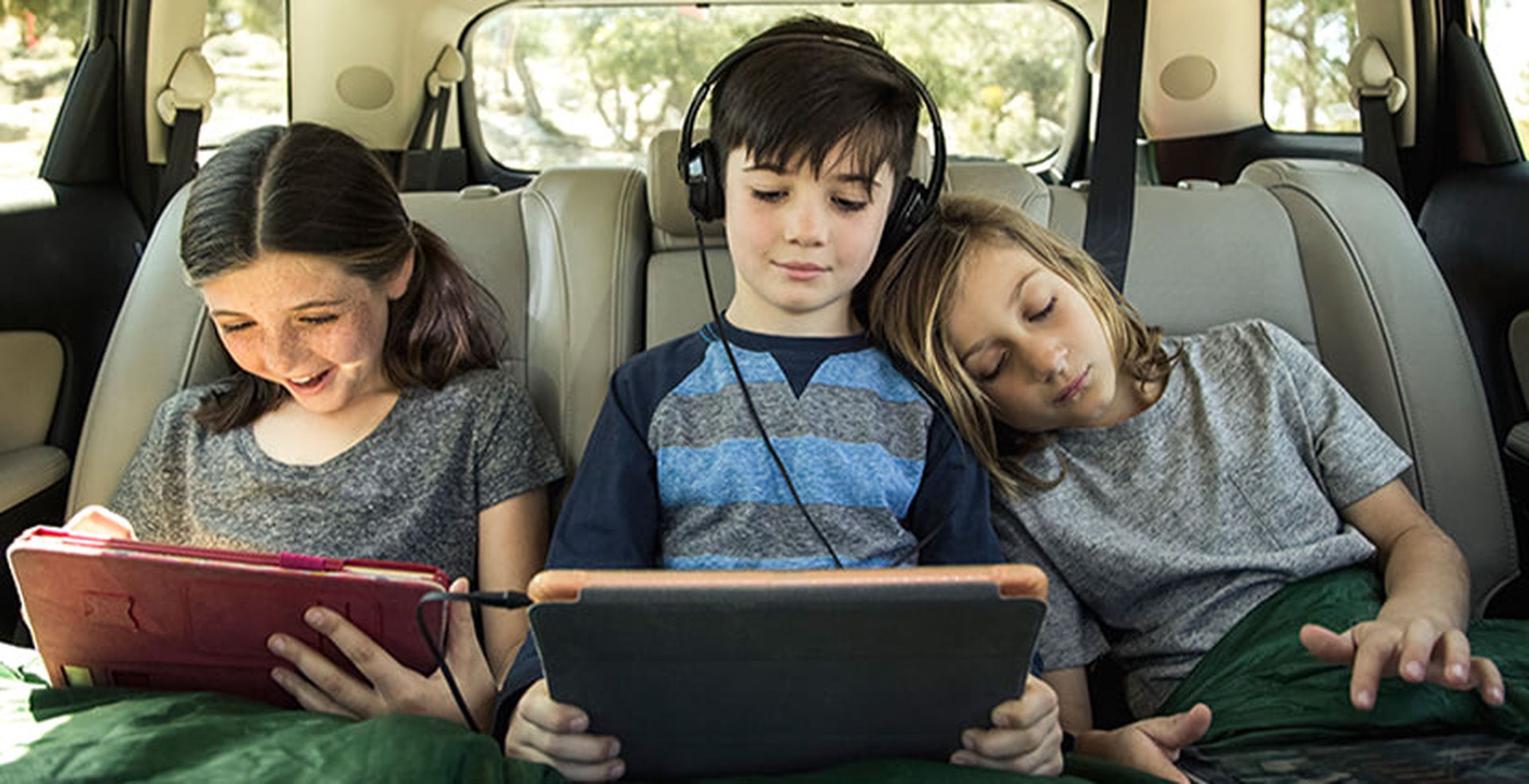 Three children in a car's backseat, engrossed in tablets; one wears headphones, another smiles, and the third leans over while watching.