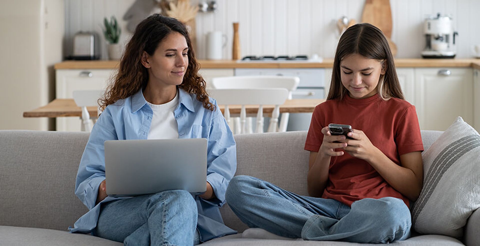 Woman with a laptop and a girl with a smartphone sit on a sofa in a cozy kitchen, both engrossed in their screens.