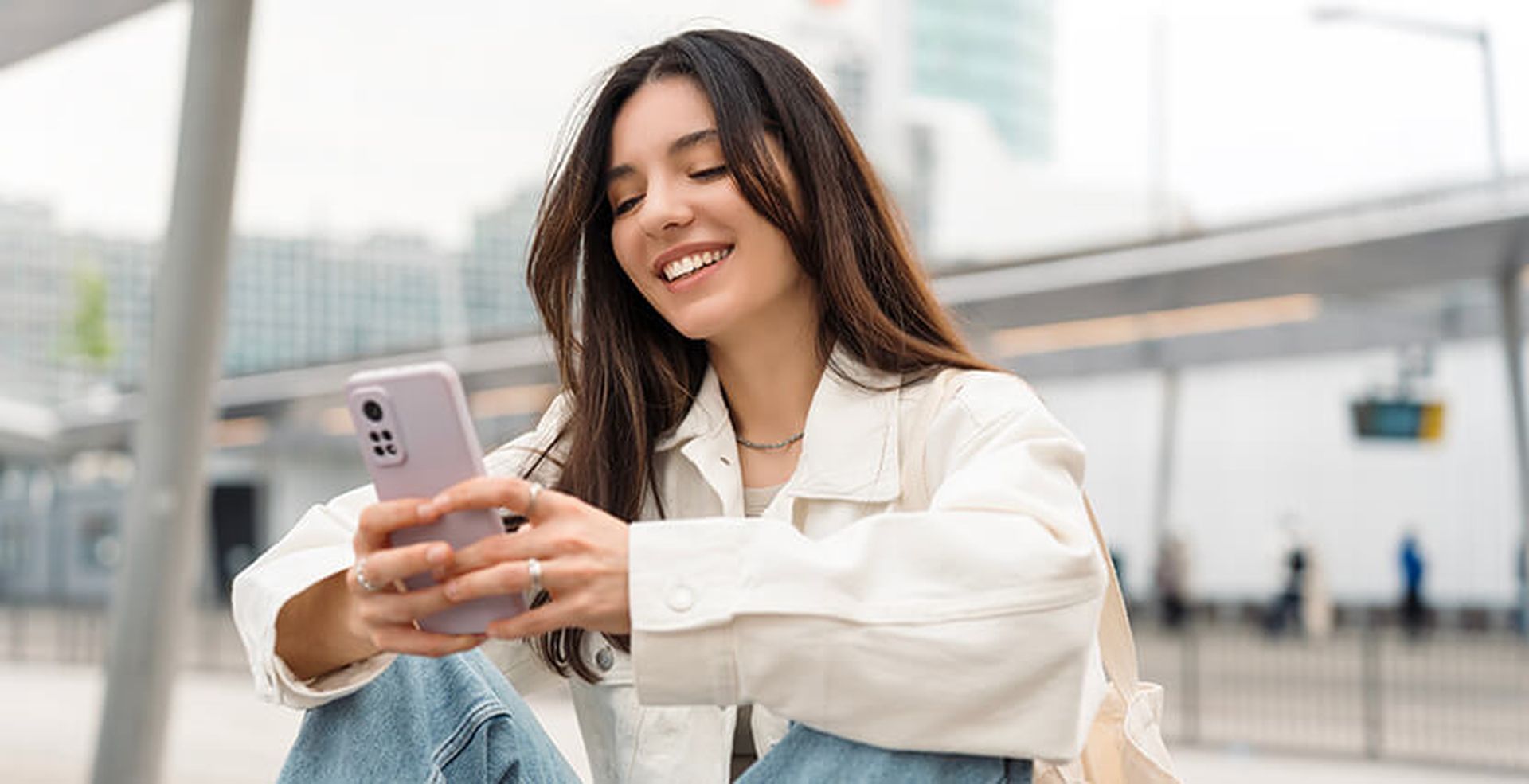 Smiling woman in a white jacket sitting outdoors, looking at her smartphone. Urban environment with blurred buildings in the background.