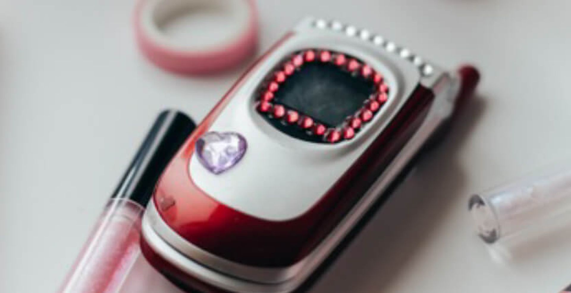 A red flip phone decorated with pink gems and a purple heart sticker, surrounded by lip gloss and tape on a white surface.