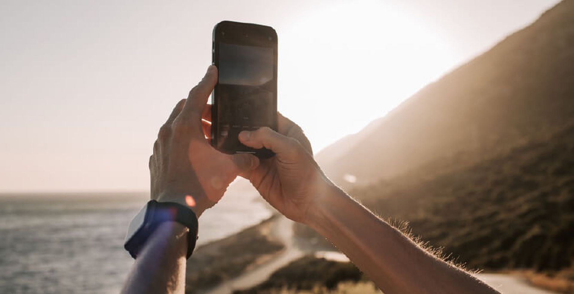 Person taking a photo with a smartphone towards the sun near a coastal hillside.