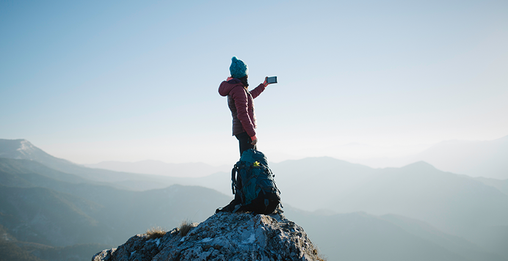 A person in winter clothing takes a selfie on a mountain peak, with a backpack beside them and a vast mountain landscape in the background.