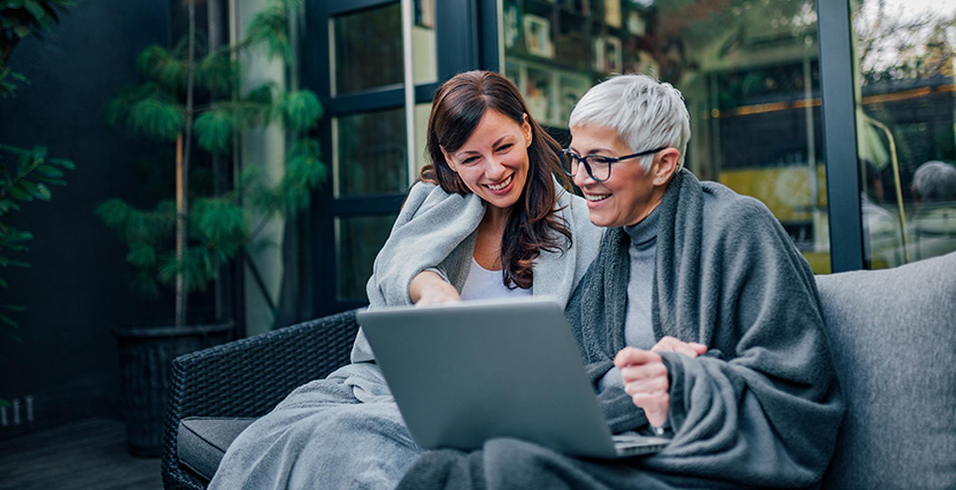 Two women, wrapped in blankets, sit on a sofa outdoors, smiling and looking at a laptop screen.
