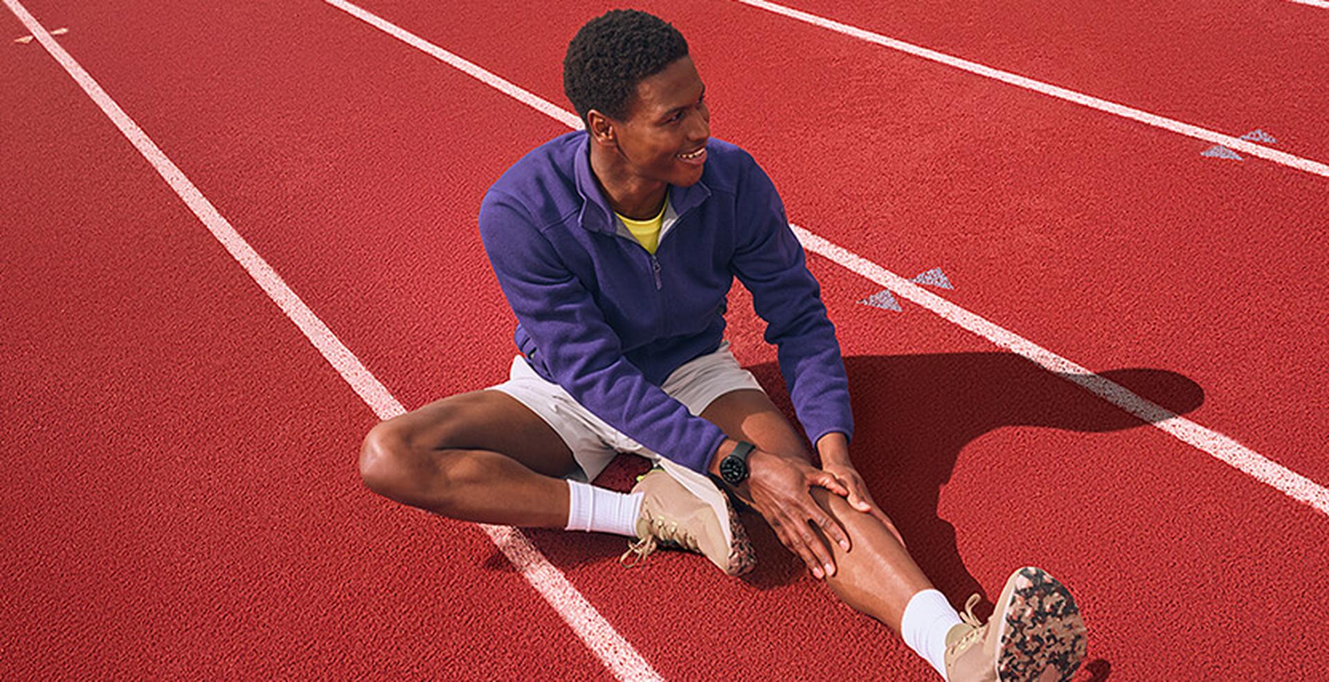 Athlete sitting on a red track, wearing a purple jacket and white shorts, stretching one leg with a smile.