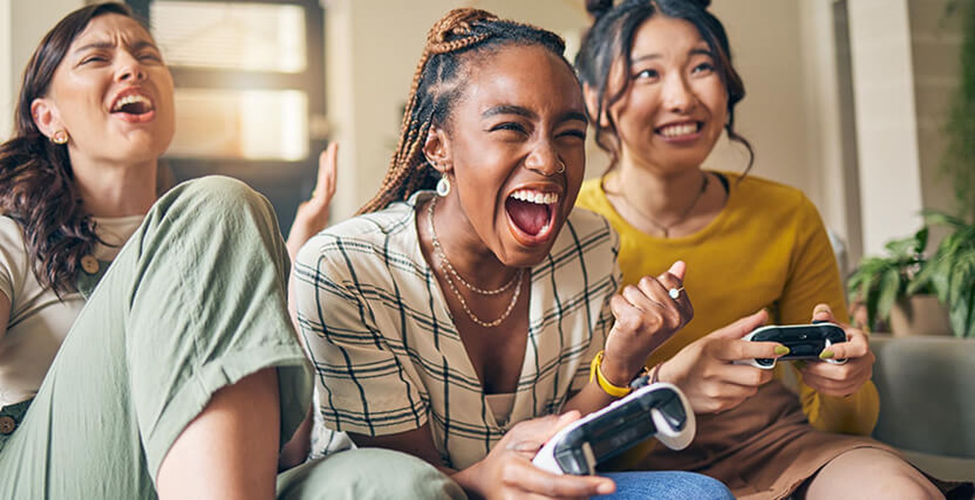 Three friends enthusiastically playing video games together on a couch, smiling and shouting, holding controllers in a lively home setting.