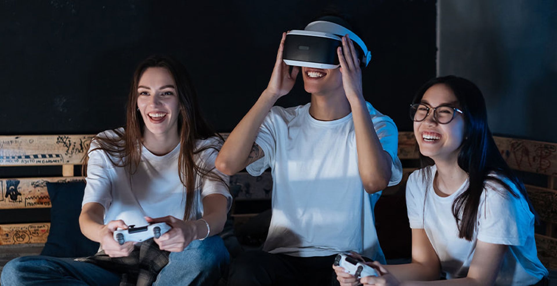 Three people sitting on a sofa playing video games; one wears a VR headset and the others hold controllers, all smiling and having fun.