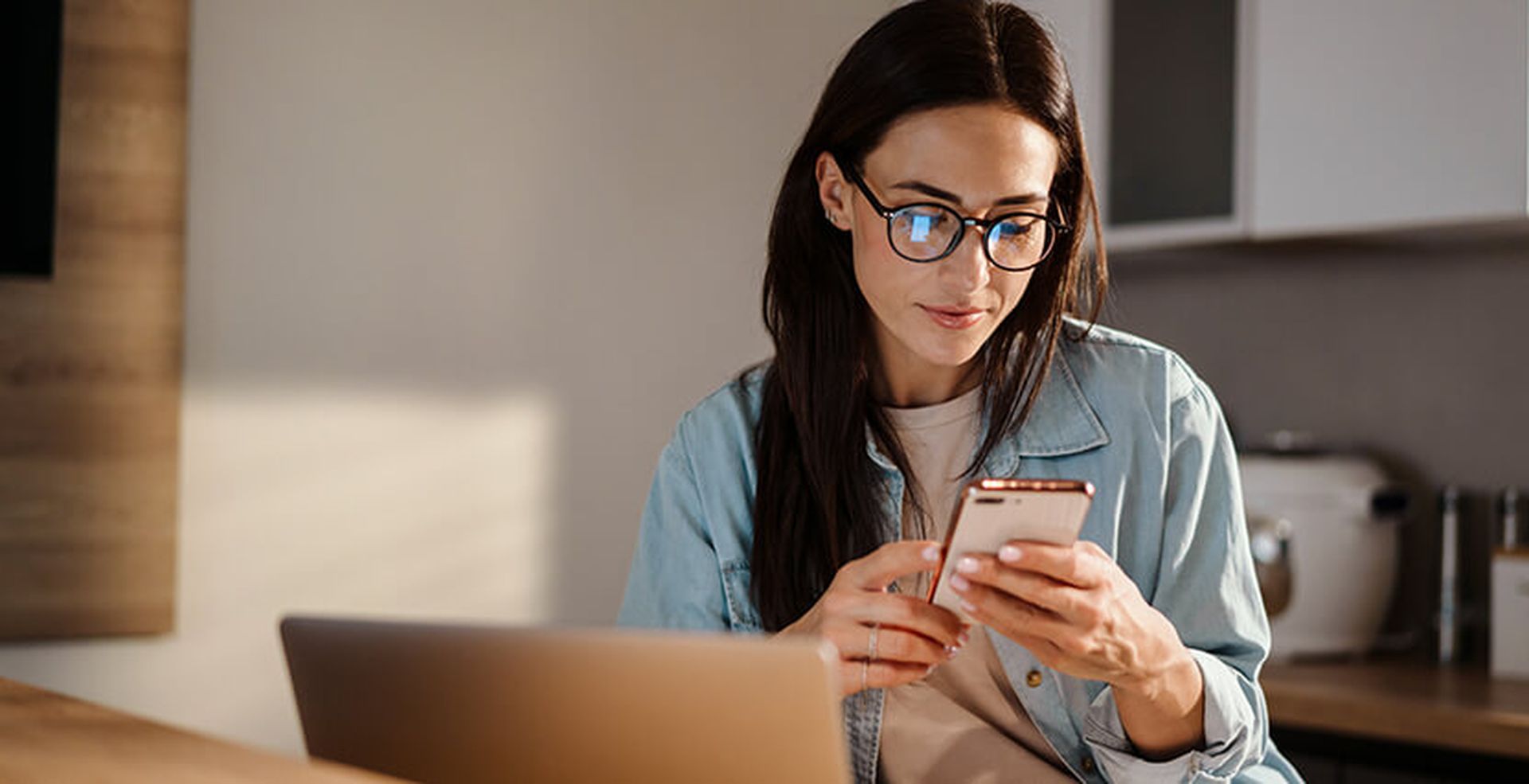 Woman with glasses using a smartphone, sitting at a table with a laptop in a cozy room.