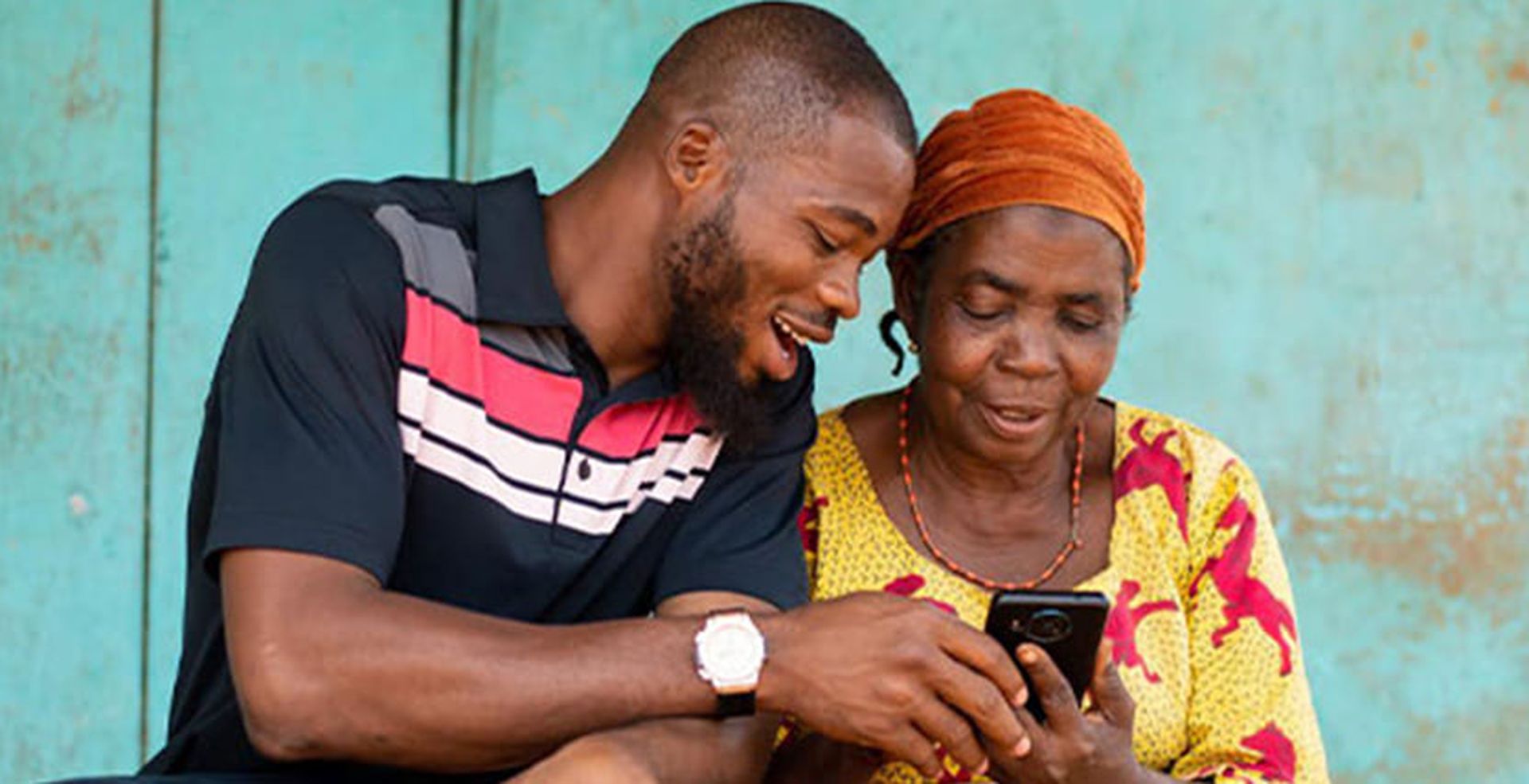 A man and an older woman sit together, smiling and looking at a smartphone in the woman's hand, set against a teal background.