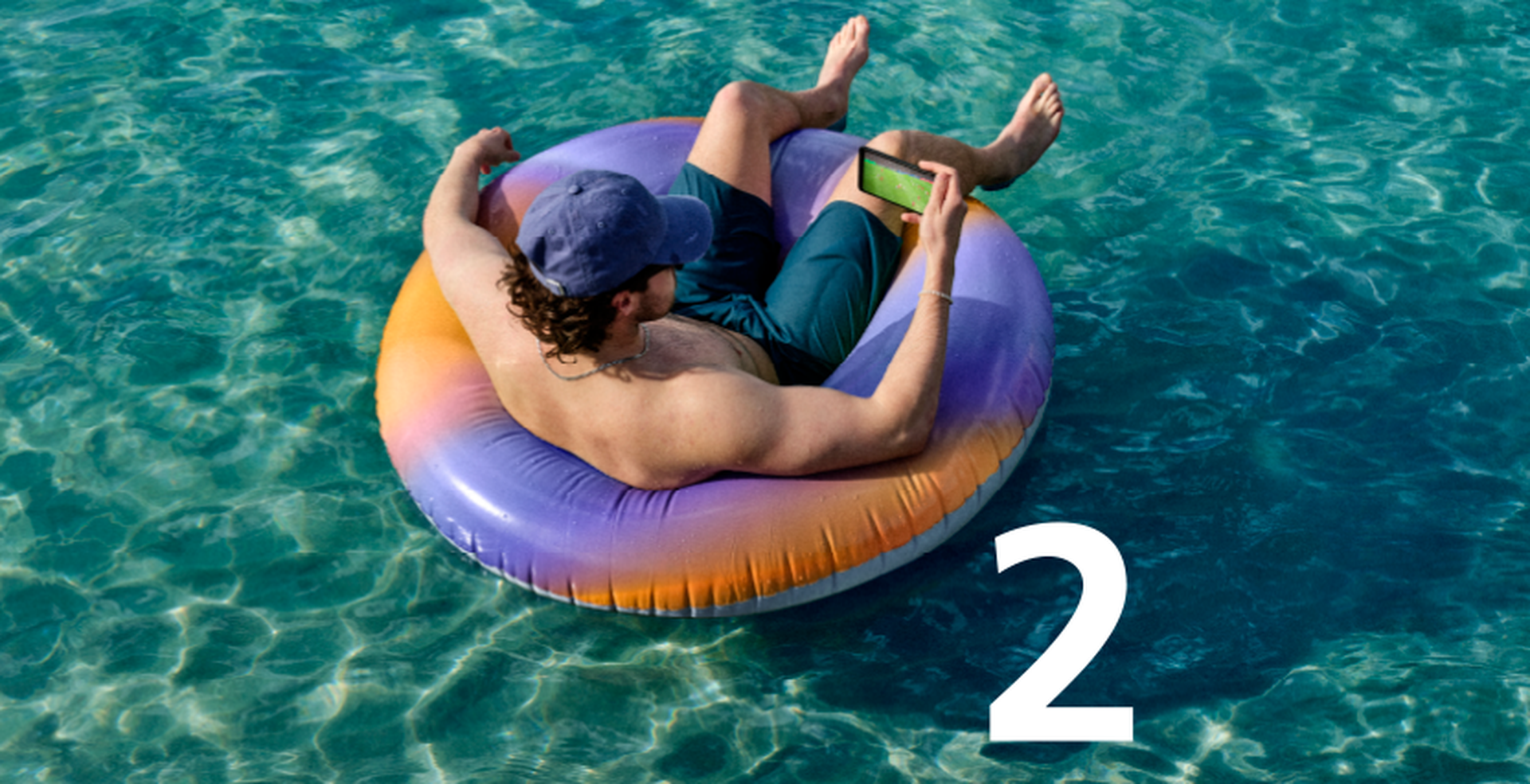 Man in a cap relaxes on a colorful inflatable ring in clear blue water, using a smartphone. Large number "2" is visible on the image.