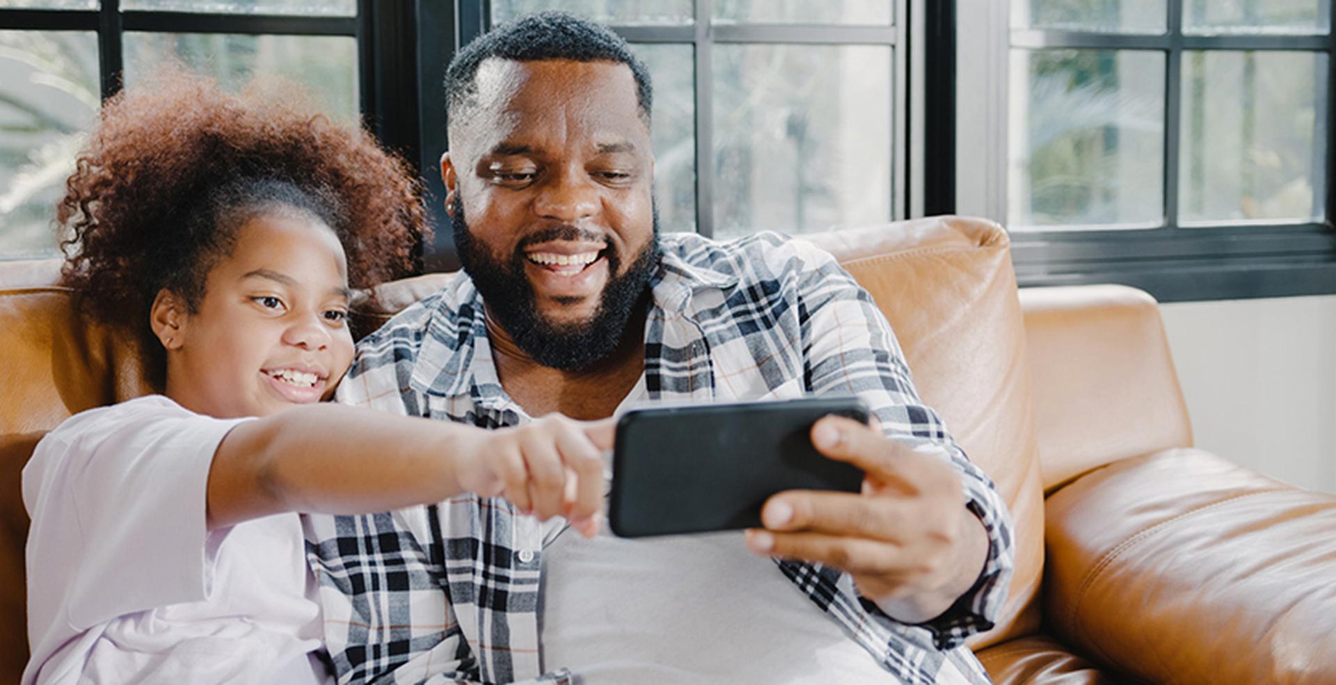 A man and a child sit on a couch, smiling and taking a selfie together with a smartphone in a bright, sunlit room.