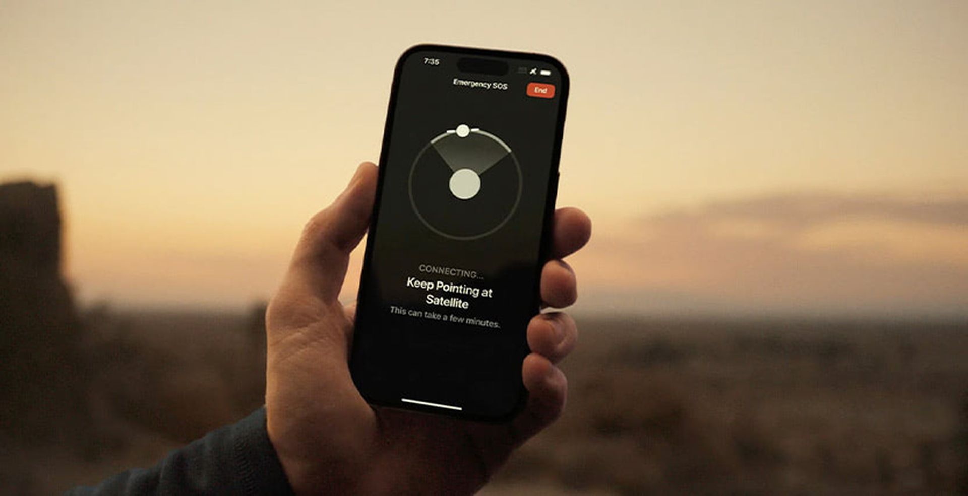 A person holds a smartphone displaying an emergency SOS satellite connection interface against a desert landscape at dusk.