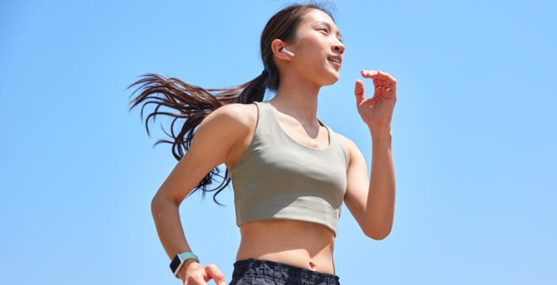 A woman in a gray sports bra running outside on a clear day, wearing wireless earbuds and a fitness tracker.