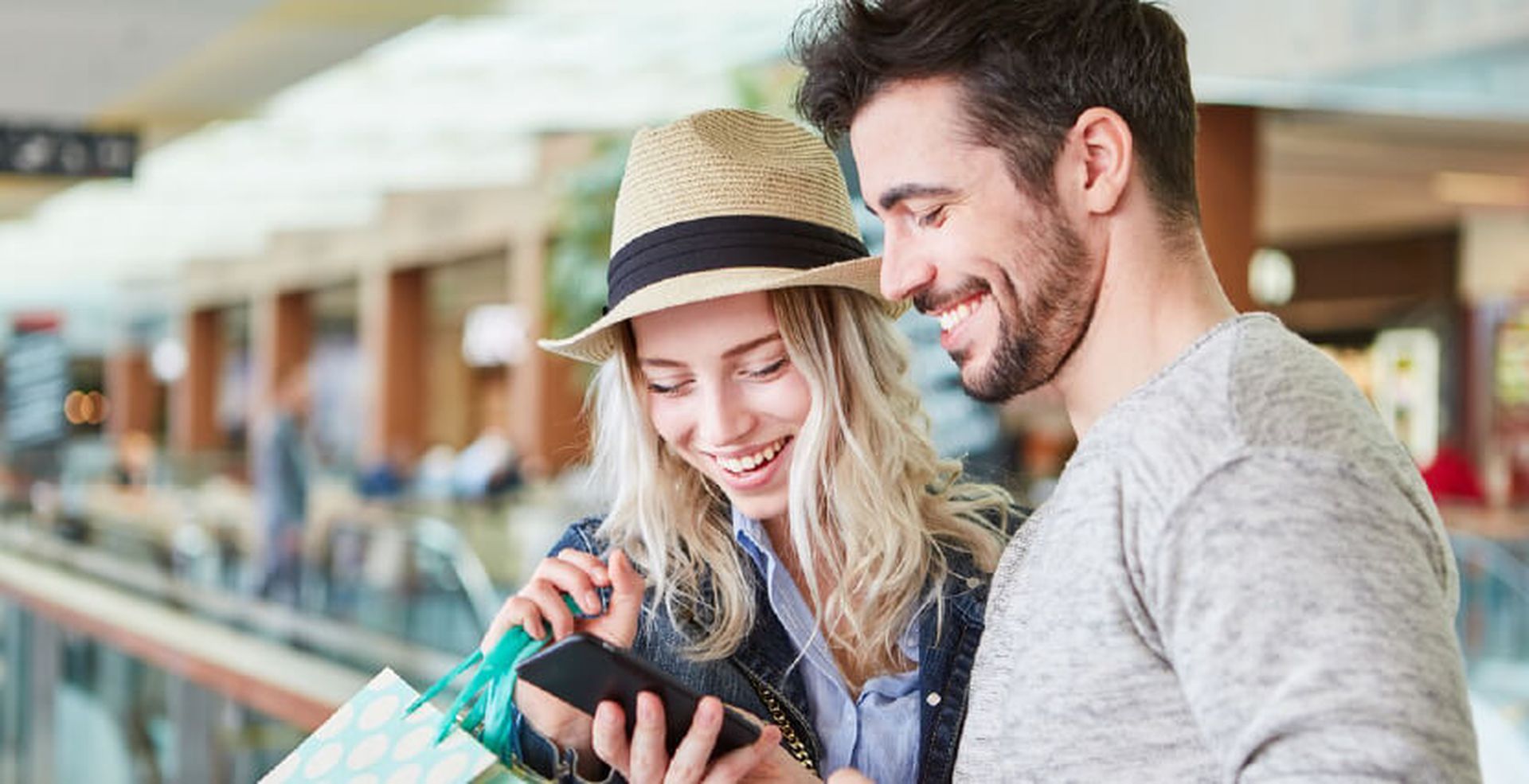 A smiling couple at a mall looks at a phone together, with shopping bags in hand.