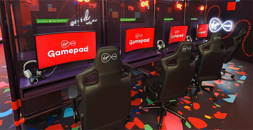 A row of gaming stations with black chairs, red screens displaying "Gamepad," and headsets, set in a colorful, modern gaming area.