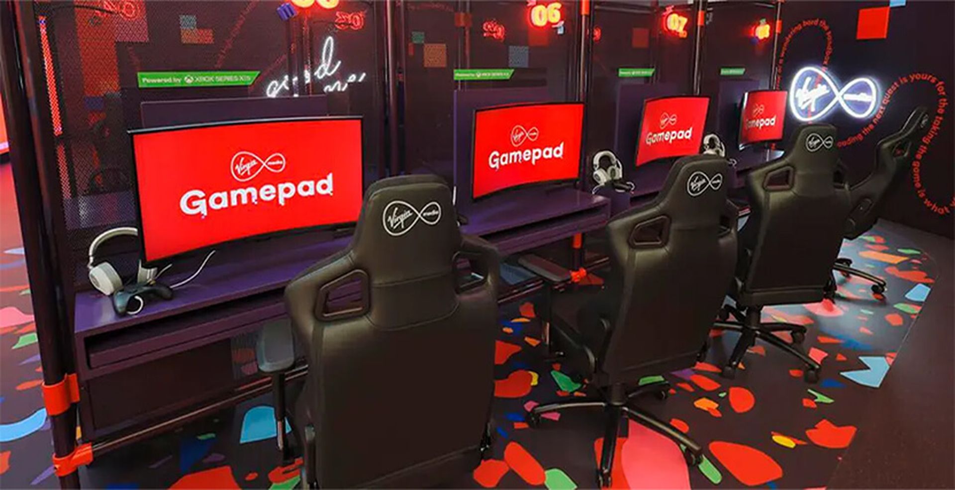 A row of gaming stations with black chairs, red screens displaying "Gamepad," and headsets, set in a colorful, modern gaming area.